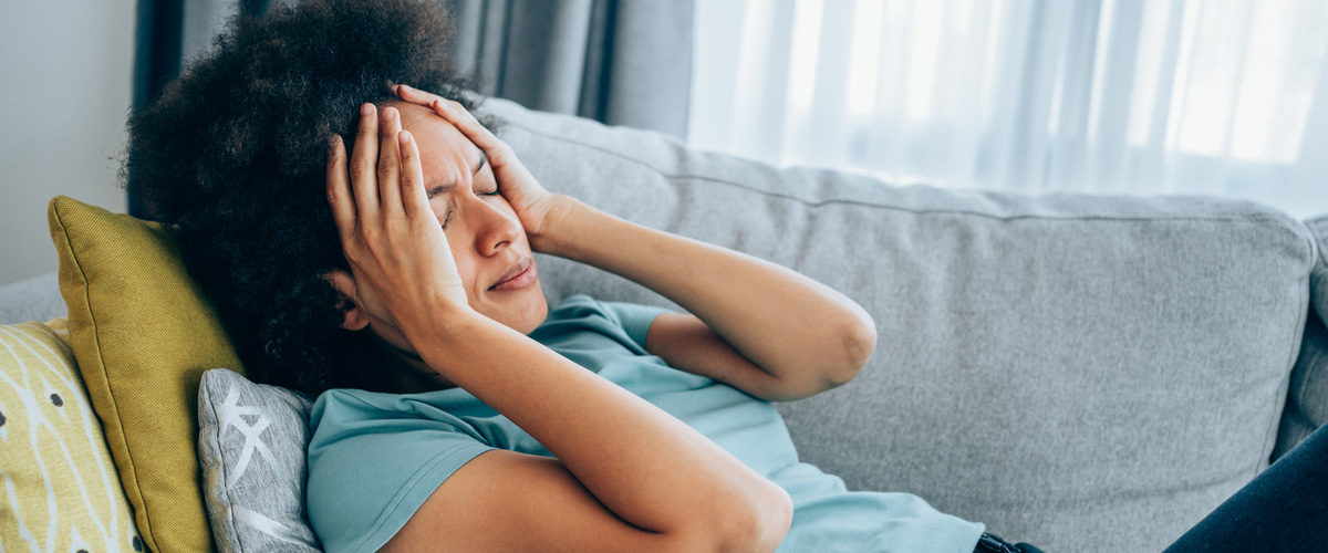 A women with curly black hair holding her head in pain while laying on a couch